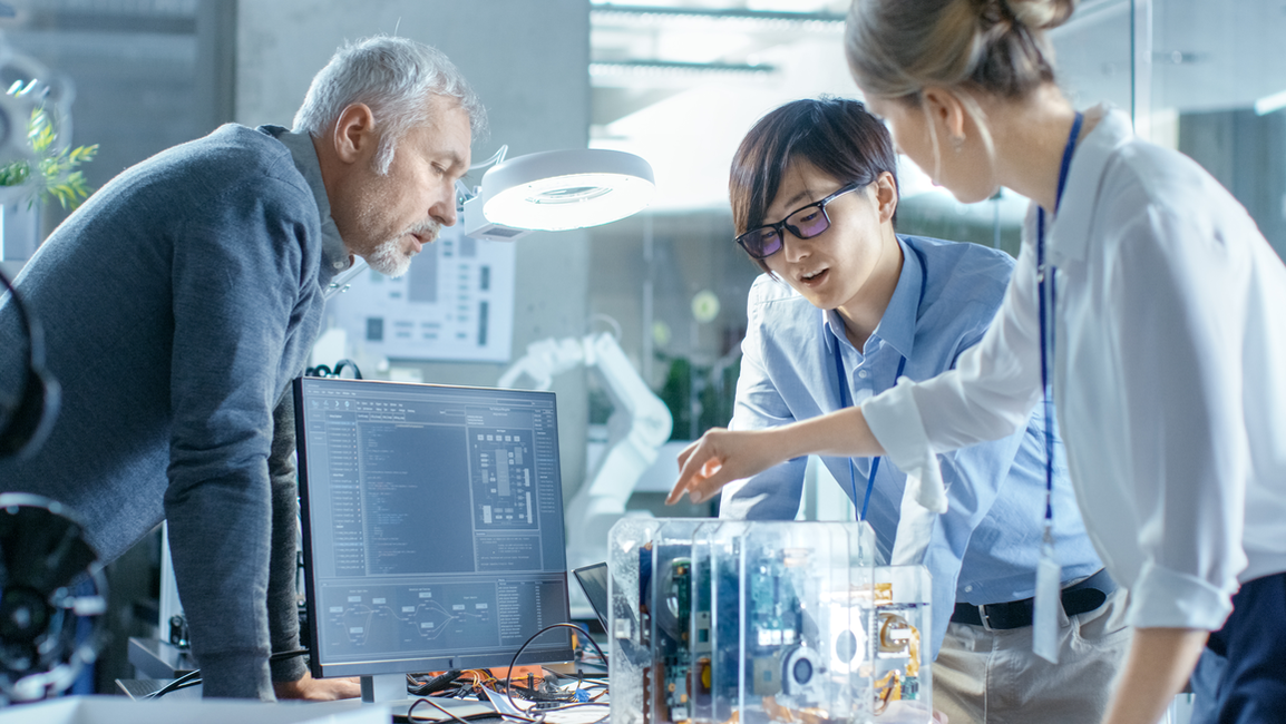 Drei Personen stehen an einem Tisch voller Technik und Computer, eine Person erklärt den anderen etwas. Atmosphäre: technisch