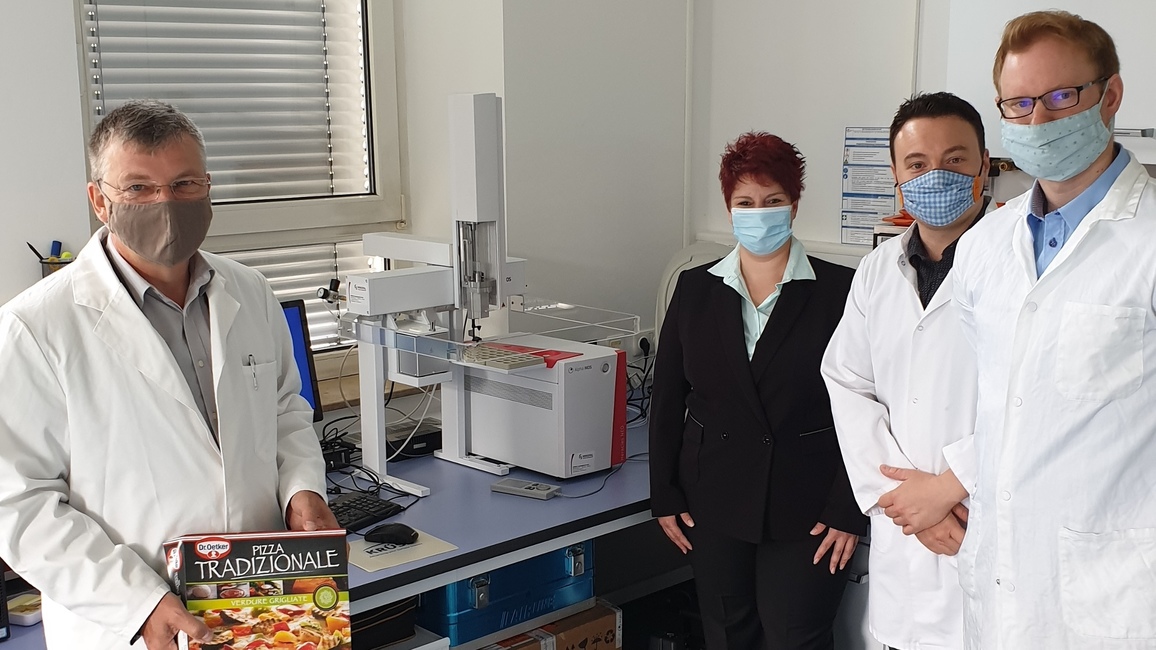 Vier Wissenschaftlerinnen und Wissenschaftler stehen in einem Labor, einer hält den Karton einer Tiefkühlpizza in der Hand