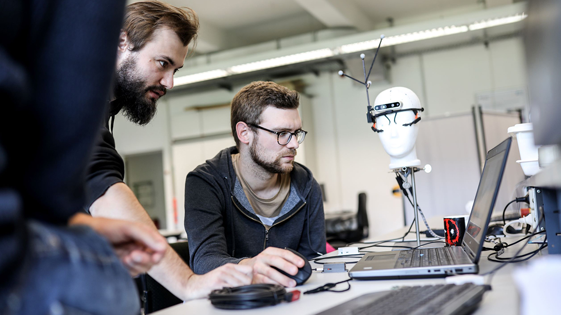 Junge Männer sitzen und stehen um einen Laptop herum; im Hintergrund ein Styroporkopf mit einem Gestell aus Kameras und Antennen