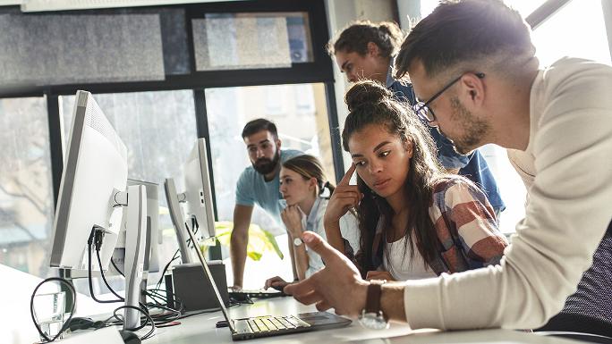 Junge Menschen sitzen und stehen an Laptops und Monitoren und diskutieren