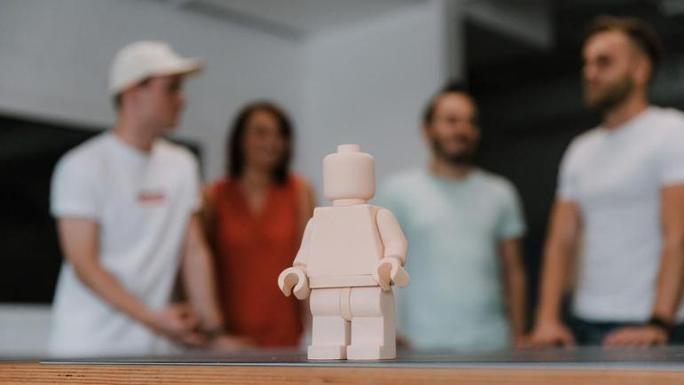Das Bild zeigt im Vordergrund ein hautfarbenes Lego-Männchen ohne Gesicht auf einem Tisch und im Hintergrund verschwommen einige junge Menschen