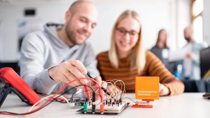 Ein junger Mann und eine junge Frau sitzen vor einem elektronischen Aufbau und arbeiten an den Kabeln und Steckern