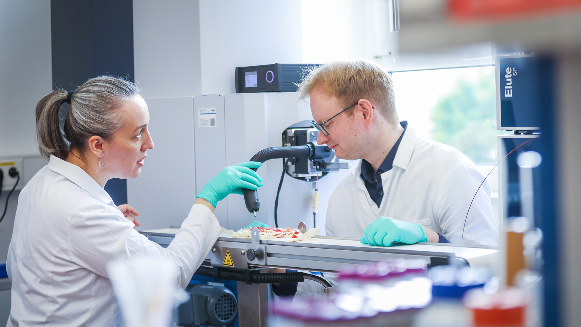 A female and a male scientist examine a frozen pizza on a production line