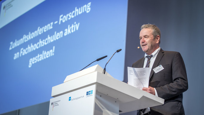 A man is standing at a speaker's desk, with a big screen in the background displaying the conference title.