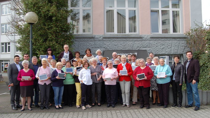 Group image of around 30 senior citizens in front of a house, many holding tablets
