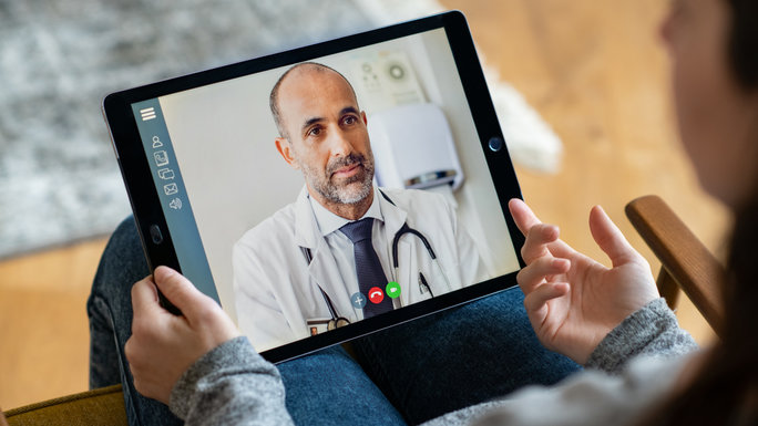 Two hands holding a tablet, the screen shows a doctor in video chat