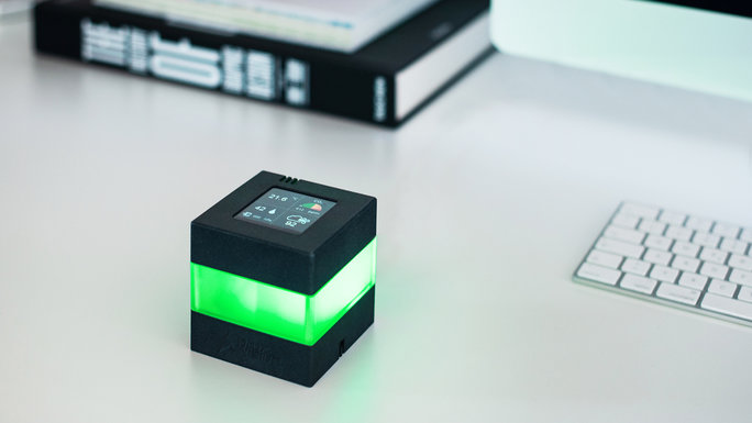 A cube-shaped device that glows in color, on a desk next to a keyboard and a folder