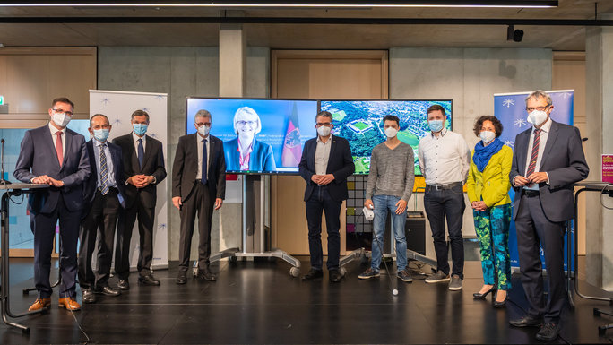 Several people wearing masks stand in a semicircle, in the middle of which is a large screen on which Minister Karliczek is connected by video