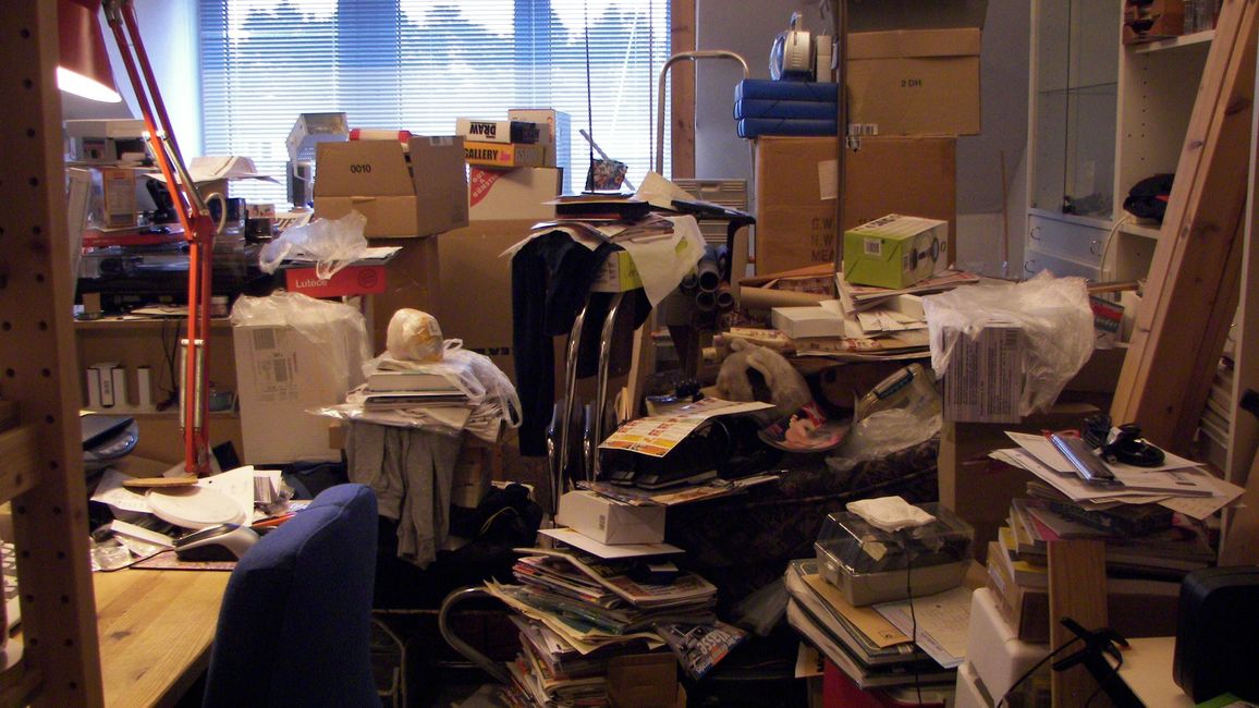 A room, filled with furniture, cardboard boxes and paper stacks. There is a desk in the front and a window in the background.