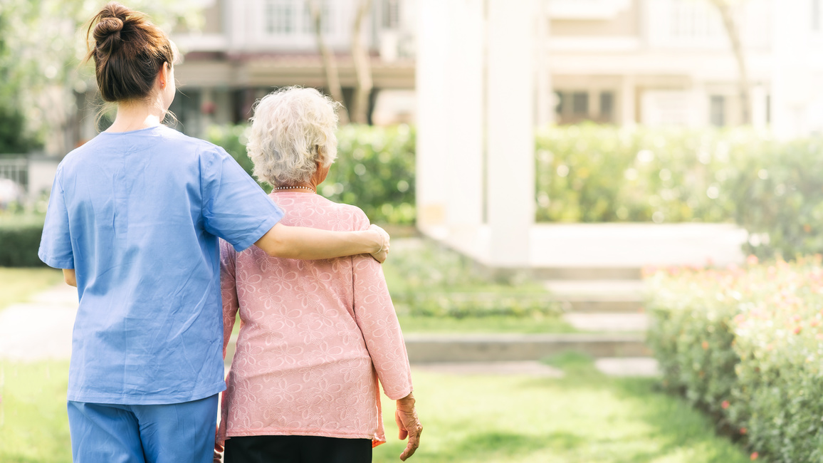 A nurse in work clothes puts her arm around an old lady. Both are photographed from behind and are standing in a park or garden