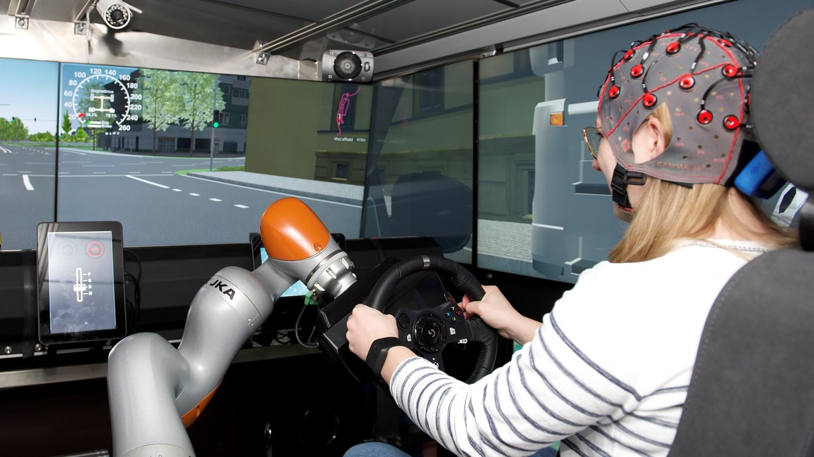 A young woman in a car seat with an EEG cap is holding the steering wheel and looks at the screen of the driving simulator