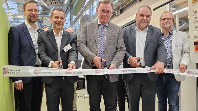 Five men are standing in a row. The three in the middle are holding scissors and are cutting a ribbon.