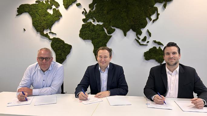 Three men are sitting at a table signing papers. A world map made of moss hangs on the wall behind them.