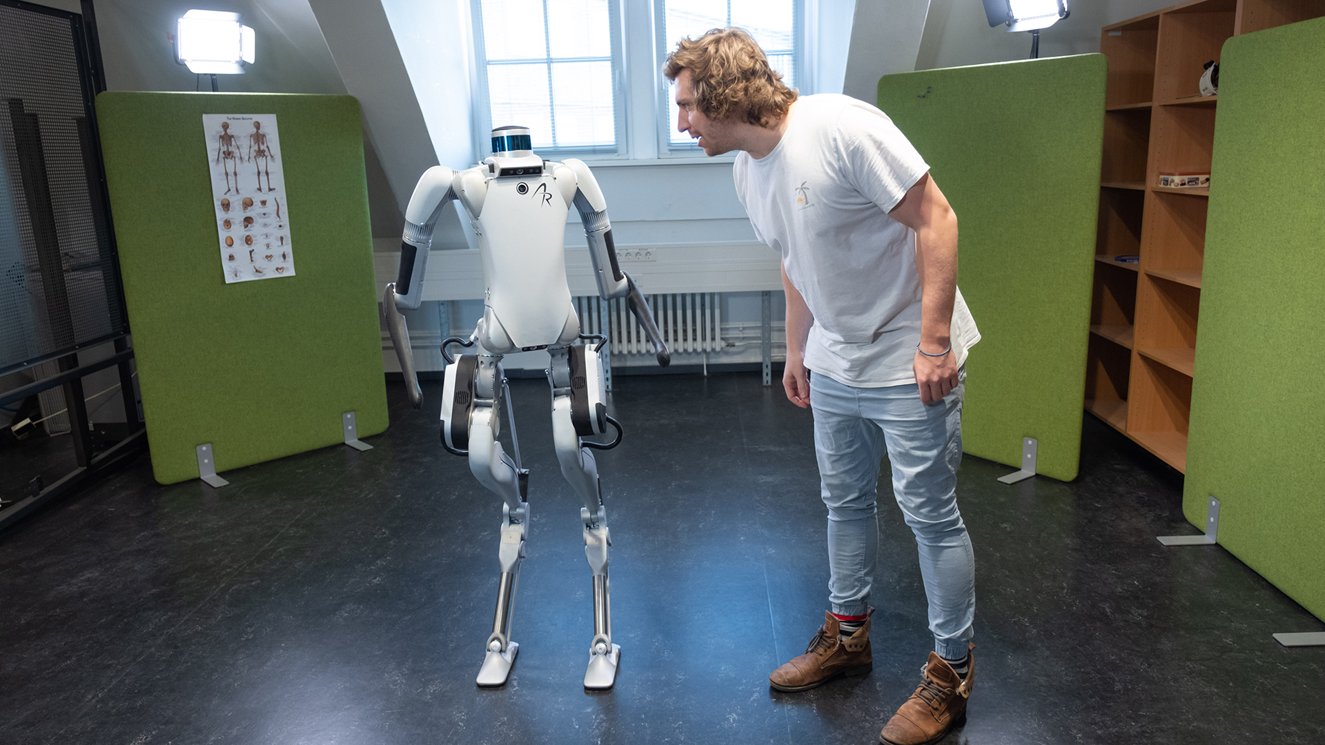 In a largely empty room, a two-legged robot and a human stand side by side and seem to be talking to each other.
