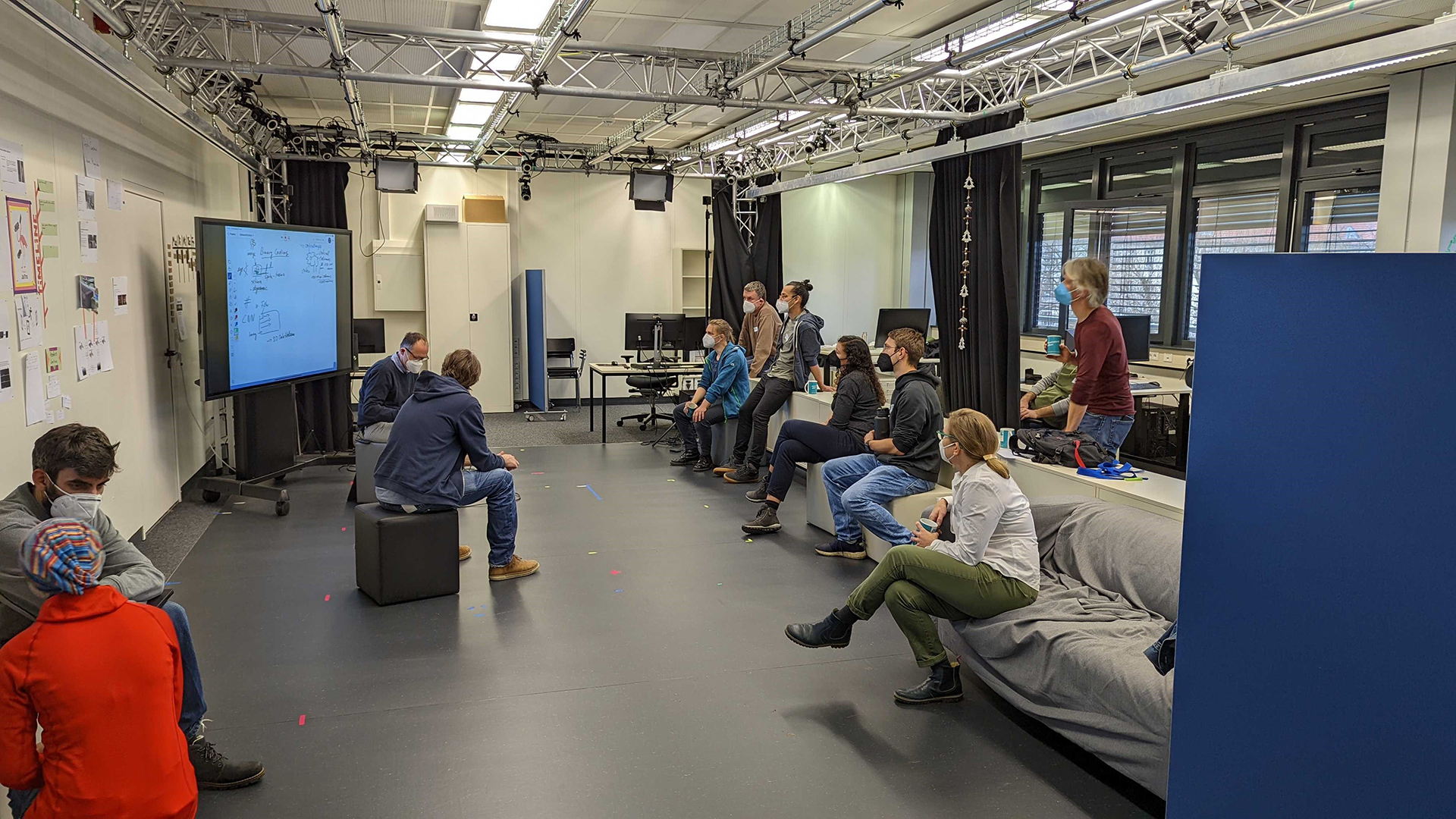About ten people sit and stand in a room, looking at a digital whiteboard and discussing with each other.