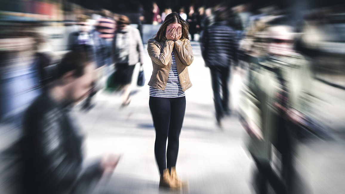 A young woman stands in the middle of people and holds her hands in front of her face in panic.