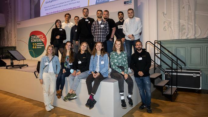 Group photo; 14 people standing and sitting next to or behind each other on one corner of the stage